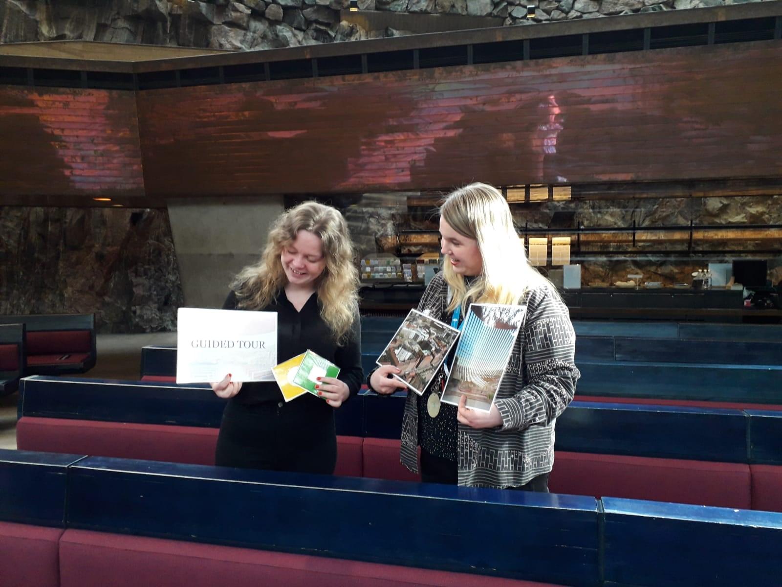 Kuvassa kaksi naista, joilla on kädessä kuvakylttejä Temppeliaukion kirkosta ja kyltti, jossa lukee 
