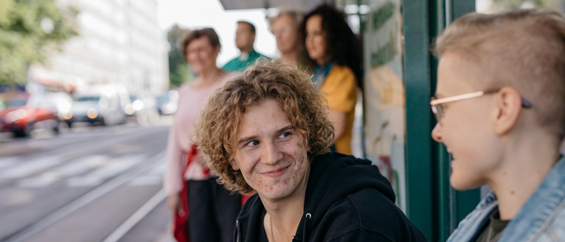 Mies ja nainen istuvat ratikkapysäkin penkillä ja katsovat toisiaan, taustalla seisoo neljä muuta ihmistä